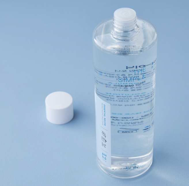 A clear, cylindrical bottle of calming toner with a white screw cap placed beside it on a light blue background. The toner bottle has blue and white text printed on it.